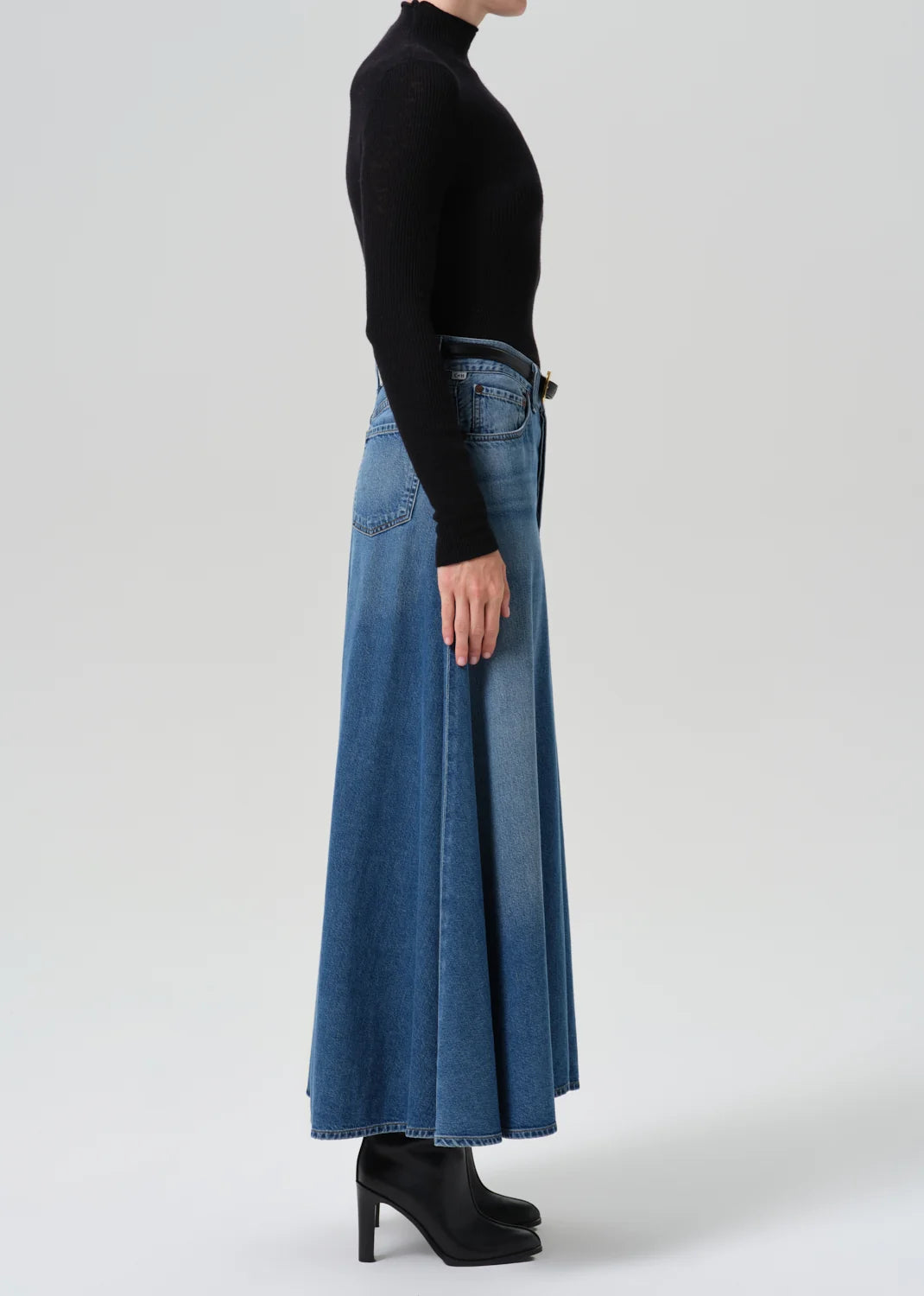 A model shown from the side wearing a black long-sleeve mock neck top, a high-waisted wide-leg blue denim skirt, and black high-heeled boots, set against a plain light gray studio background.