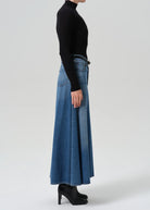 A model shown from the side wearing a black long-sleeve mock neck top, a high-waisted wide-leg blue denim skirt, and black high-heeled boots, set against a plain light gray studio background.