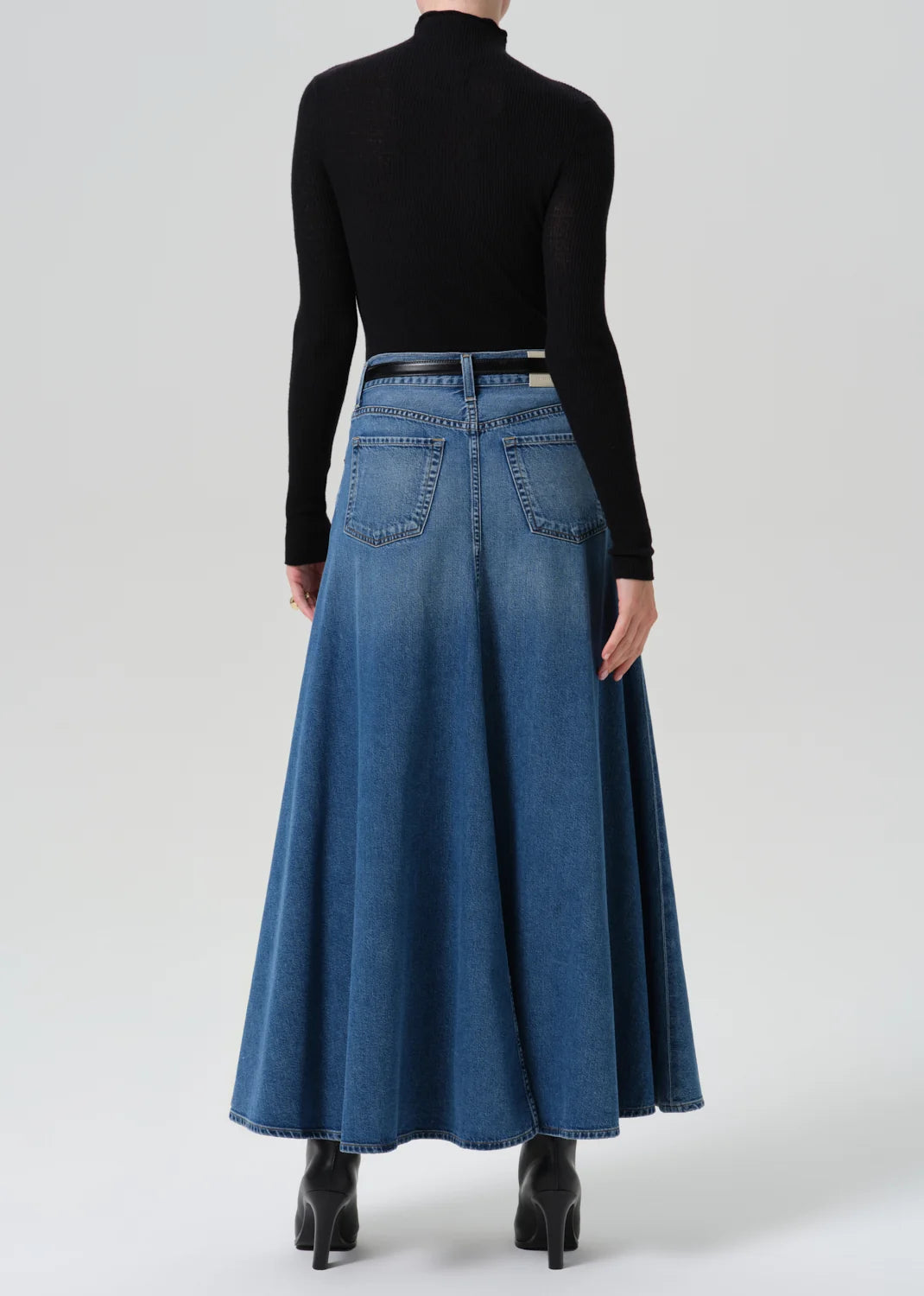 A model shown from the back wearing a long-sleeve black mock neck top and a high-waisted, flared blue denim maxi skirt with a thin black belt, paired with black ankle boots, against a light gray studio background.