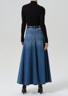 A model shown from the back wearing a long-sleeve black mock neck top and a high-waisted, flared blue denim maxi skirt with a thin black belt, paired with black ankle boots, against a light gray studio background.