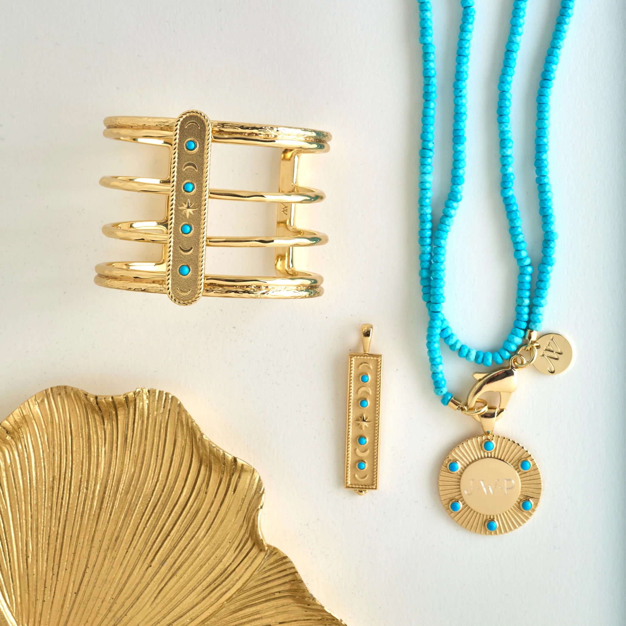 Gold jewelry set featuring a wide cuff bracelet, a vertical bar pendant, a round medallion pendant, and a turquoise beaded necklace, arranged on a white background with a gold leaf prop.