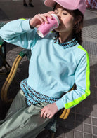 Person wearing a light blue sweater with green sleeves, drinking from a pink cup outdoors.