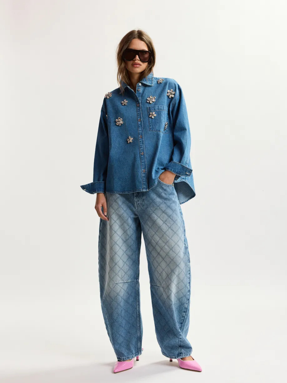 A model wearing a blue denim button-up shirt with floral applique and matching wide-leg quilted denim pants, styled with pink pointed shoes and dark sunglasses against a plain white studio background.