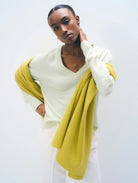 A model wearing a bright yellow-green scarf over a light green V-neck sweater and white trousers, standing against a plain white studio background.