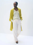 A model wearing a bright yellow-green open-front cardigan over a cream V-neck top and white wide-leg trousers, standing against a plain white studio background.