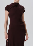 A woman wearing a dark burgundy knit dress with a draped mock neck and short cap sleeves, shown from the back against a light gray studio background.