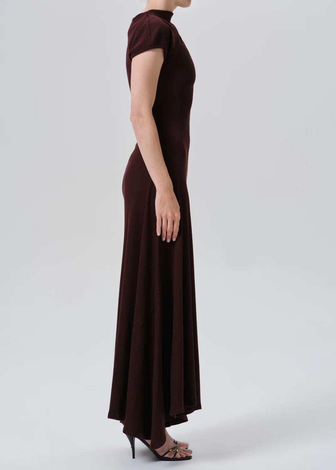 A woman modeling a dark burgundy maxi dress with short sleeves, a mock neck, and a fitted silhouette, shown in profile against a light gray studio background.