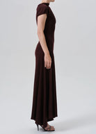 A woman modeling a dark burgundy maxi dress with short sleeves, a mock neck, and a fitted silhouette, shown in profile against a light gray studio background.