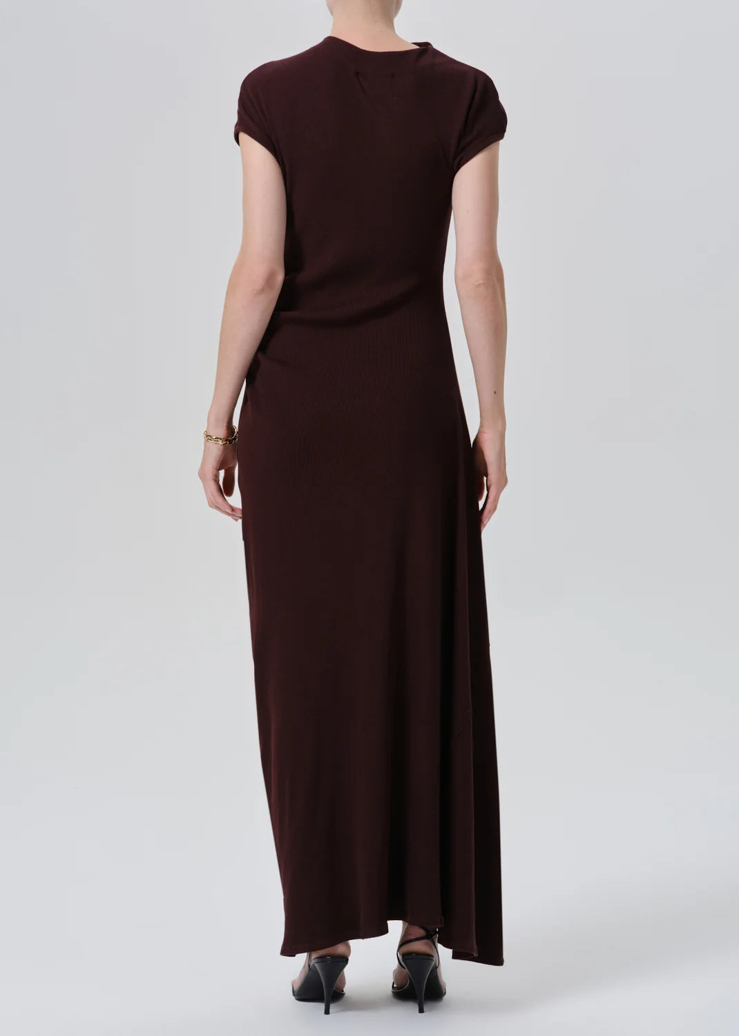 A woman viewed from the back wearing a dark brown short-sleeve maxi dress and black heels, standing against a light gray studio background.