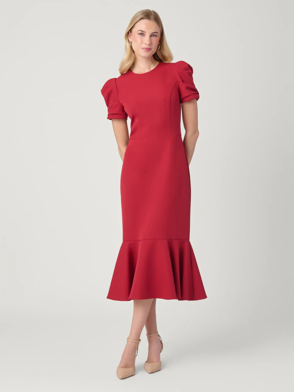 A woman modeling a red midi dress with puffed short sleeves and a flared ruffle hem, paired with nude high heels, against a light gray studio background.