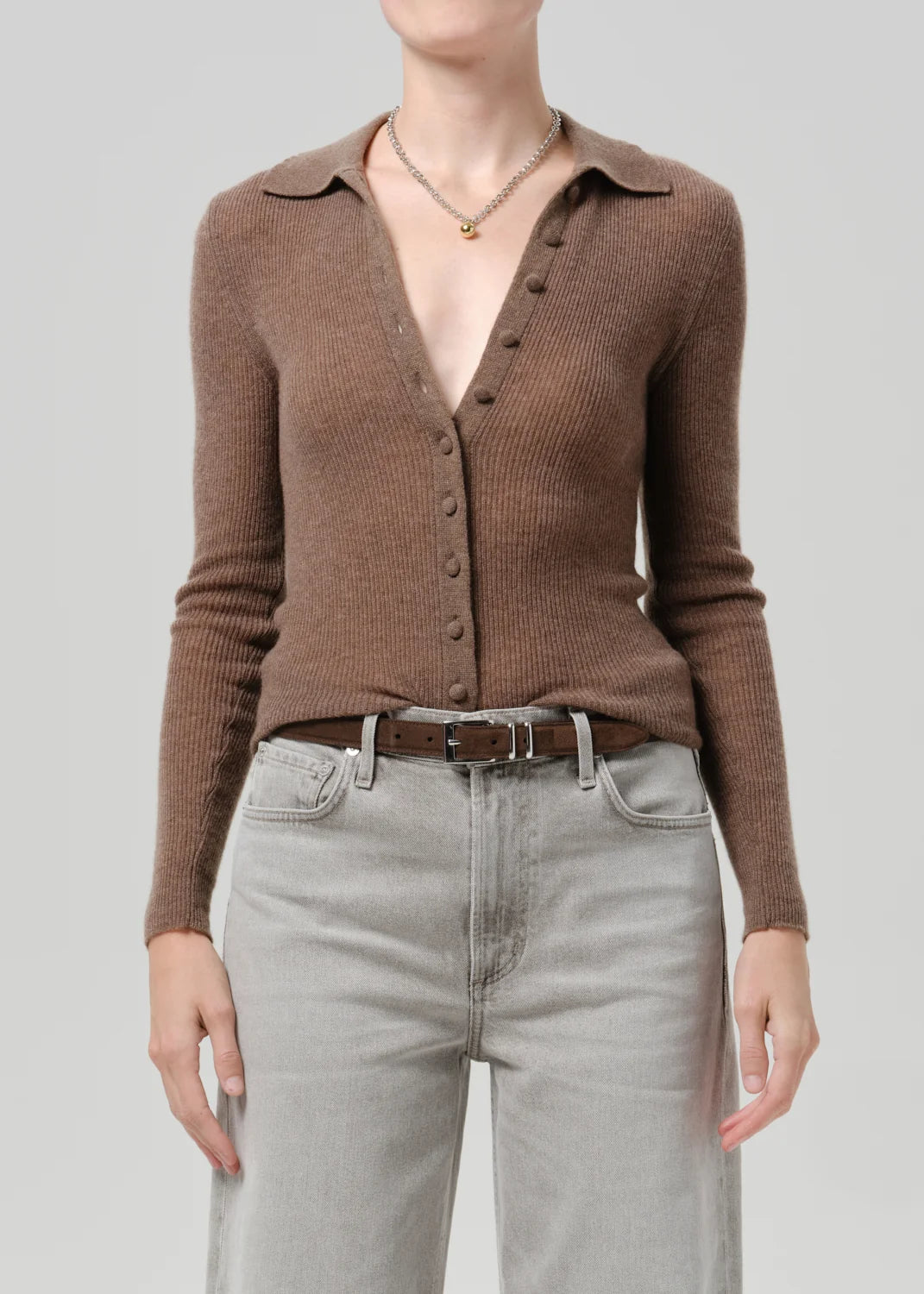 A model wearing a brown ribbed knit button-up cardigan with a collar, paired with light gray jeans and a brown belt, against a solid gray background.