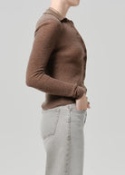 A side-profile view of a person wearing a brown ribbed knit button-front cardigan with a collar, paired with light gray jeans, against a solid light gray background.