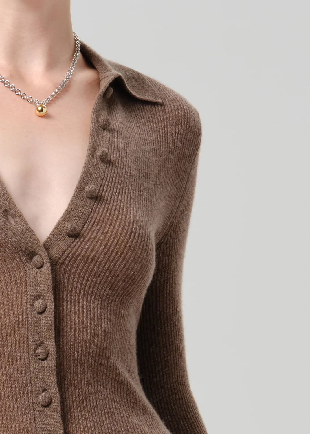 A close-up of a model wearing a brown ribbed knit button-front cardigan with a collar and a silver chain necklace with a small gold bead pendant, set against a solid light gray background.