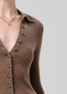 A close-up of a model wearing a brown ribbed knit button-front cardigan with a collar and a silver chain necklace with a small gold bead pendant, set against a solid light gray background.
