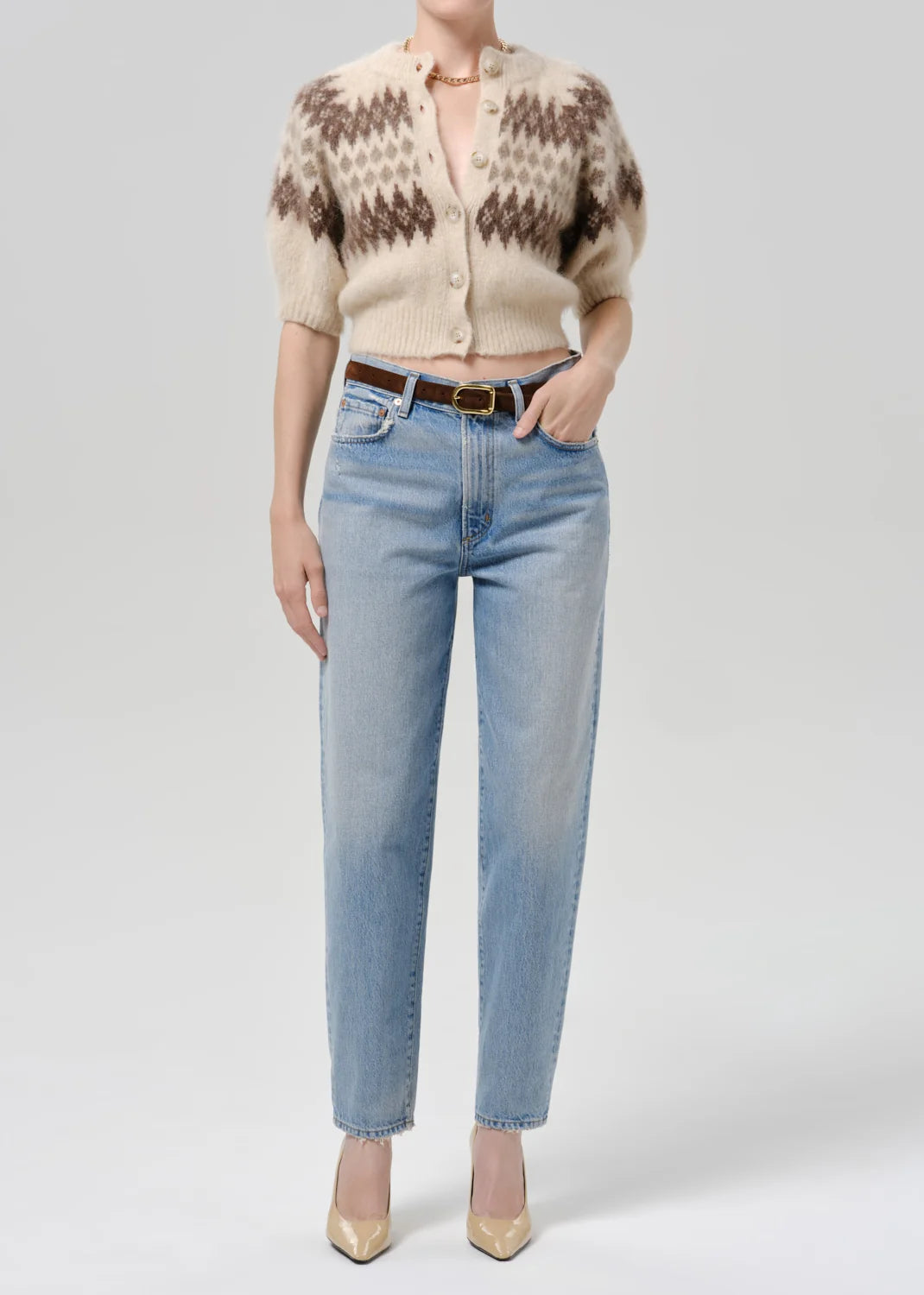 A model wearing a beige and brown patterned short-sleeve cardigan, light-wash blue jeans, a brown belt, and beige heels, photographed against a light gray studio background.