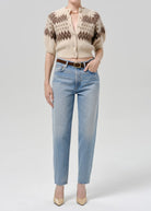 A model wearing a beige and brown patterned short-sleeve cardigan, light-wash blue jeans, a brown belt, and beige heels, photographed against a light gray studio background.