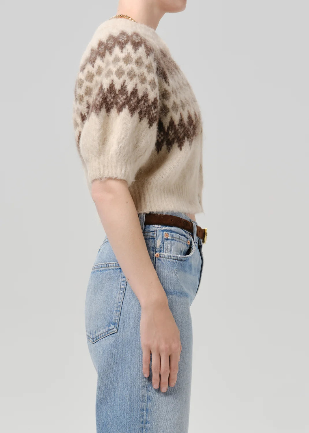 A side-profile view of a person wearing a cream-colored fuzzy short-sleeve sweater with a brown geometric pattern, paired with light blue jeans and a brown belt, against a solid light gray background.