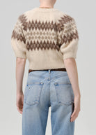 A person shown from the back wearing a cream knit sweater with a brown geometric pattern across the upper back and shoulders, paired with light blue jeans, against a plain light gray studio background.