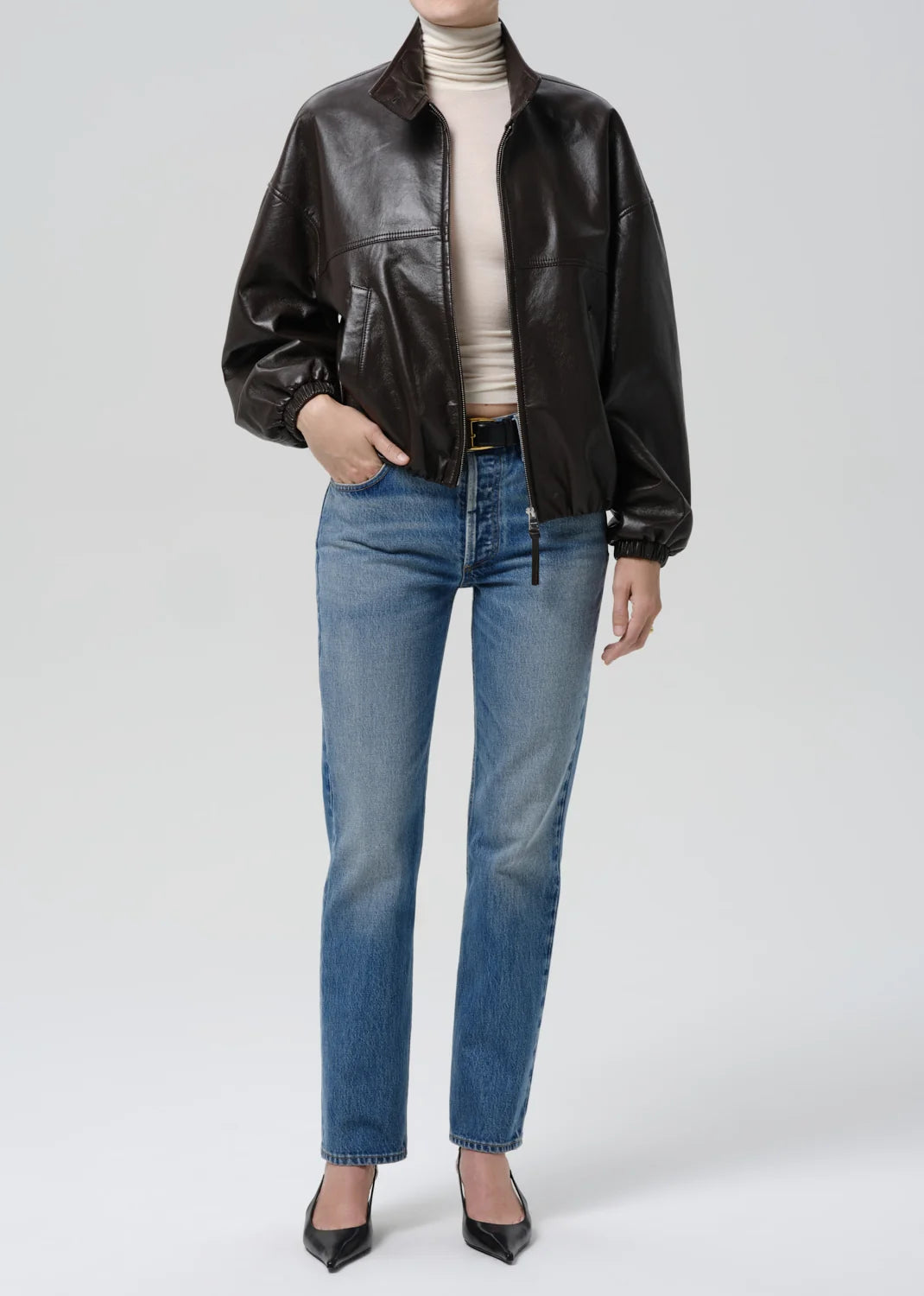 Person wearing a brown leather jacket, beige turtleneck, blue jeans, and black shoes on a white background