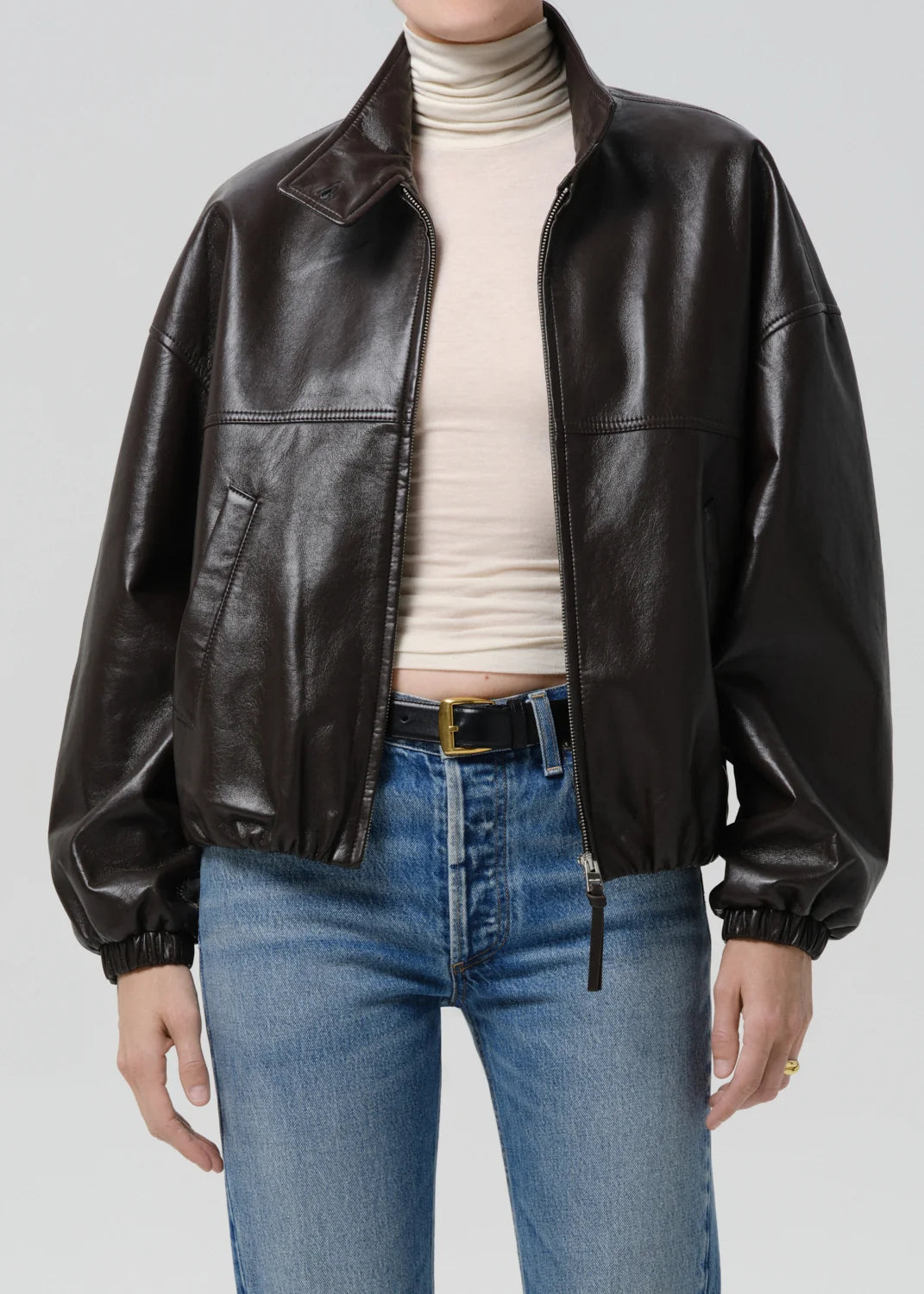 Person wearing a brown leather jacket, white turtleneck, and blue jeans on a light gray background