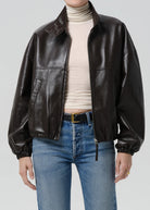 Person wearing a brown leather jacket, white turtleneck, and blue jeans on a light gray background