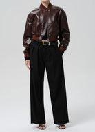 A model wearing a dark brown faux-leather bomber jacket, black wide-leg trousers, and black sandals, photographed against a neutral light gray studio background.