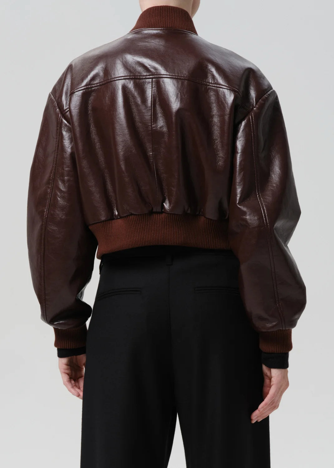 Back view of a person wearing a cropped dark brown leather bomber jacket with ribbed brown collar, cuffs, and hem, paired with black trousers, against a light gray studio background.