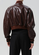 Back view of a person wearing a cropped dark brown leather bomber jacket with ribbed brown collar, cuffs, and hem, paired with black trousers, against a light gray studio background.