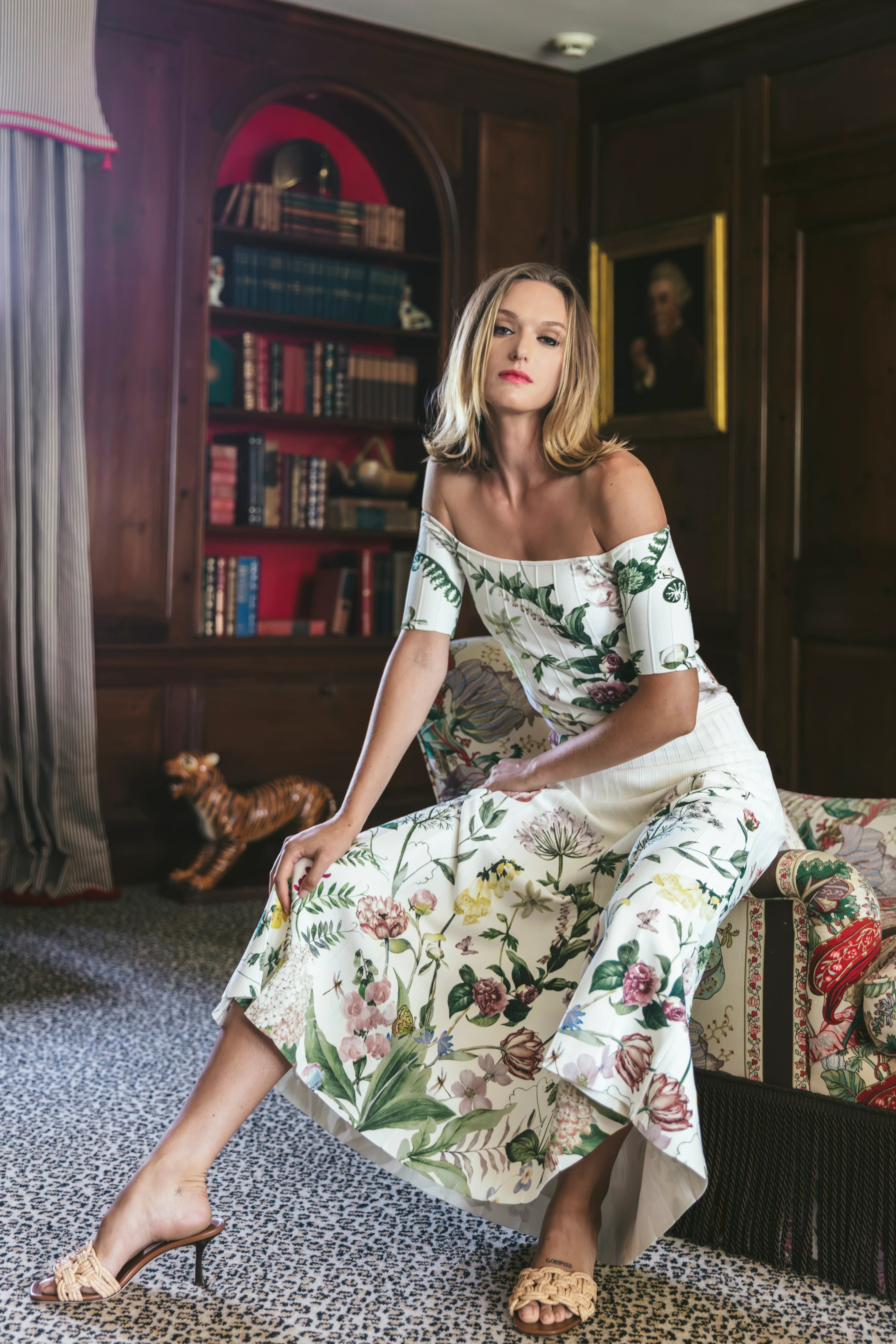 Woman in a floral dress sitting in a room with bookshelves and a dog.