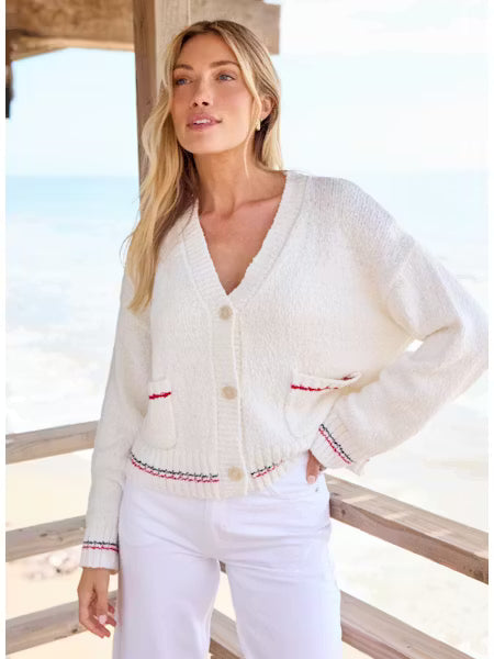 Woman wearing a white cardigan with red accents by a window with a view of water.