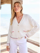 Woman wearing a white cardigan with red accents by a window with a view of water.