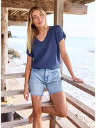 Woman in a blue top and denim shorts standing on a wooden deck by the sea.