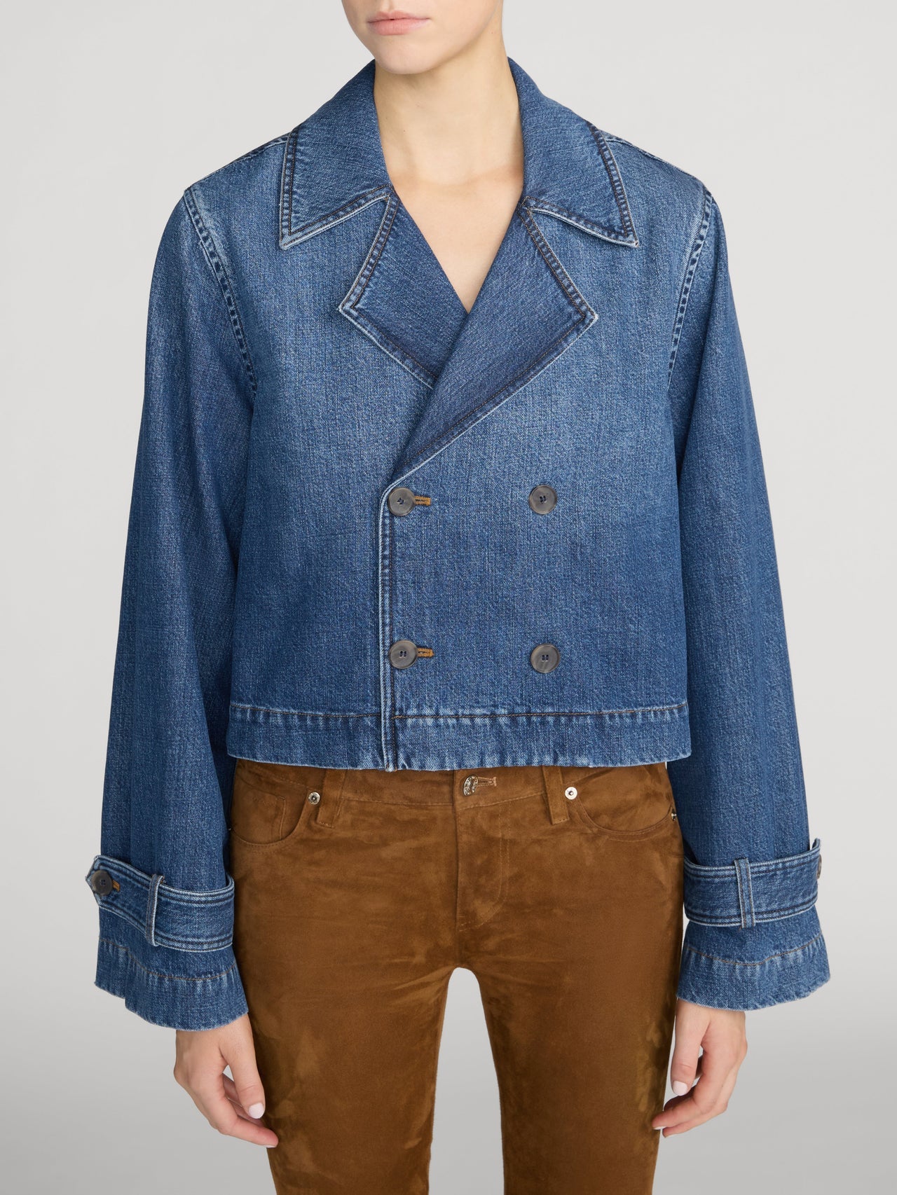 A model wearing a cropped blue denim jacket with a double-breasted design, paired with brown pants, against a light gray studio background.