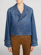 A model wearing a cropped blue denim jacket with a double-breasted design, paired with brown pants, against a light gray studio background.