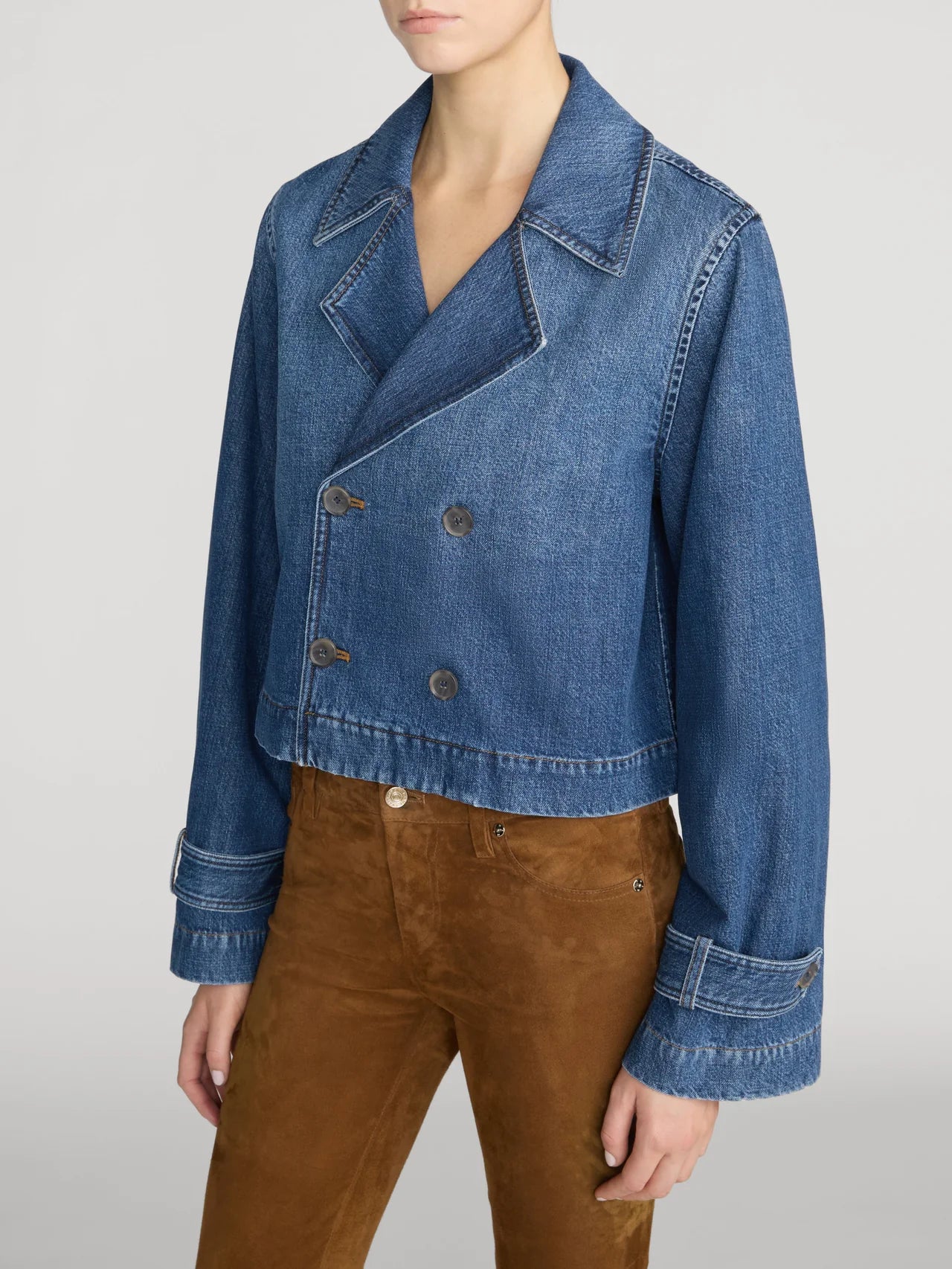 A model wearing a cropped blue denim jacket with a double-breasted button closure and brown suede pants, shown from the neck down against a light gray studio background.