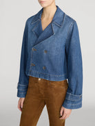 A model wearing a cropped blue denim jacket with a double-breasted button closure and brown suede pants, shown from the neck down against a light gray studio background.