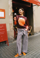 Woman wearing a maroon sweater with a pumpkin design and striped pants standing on a street.