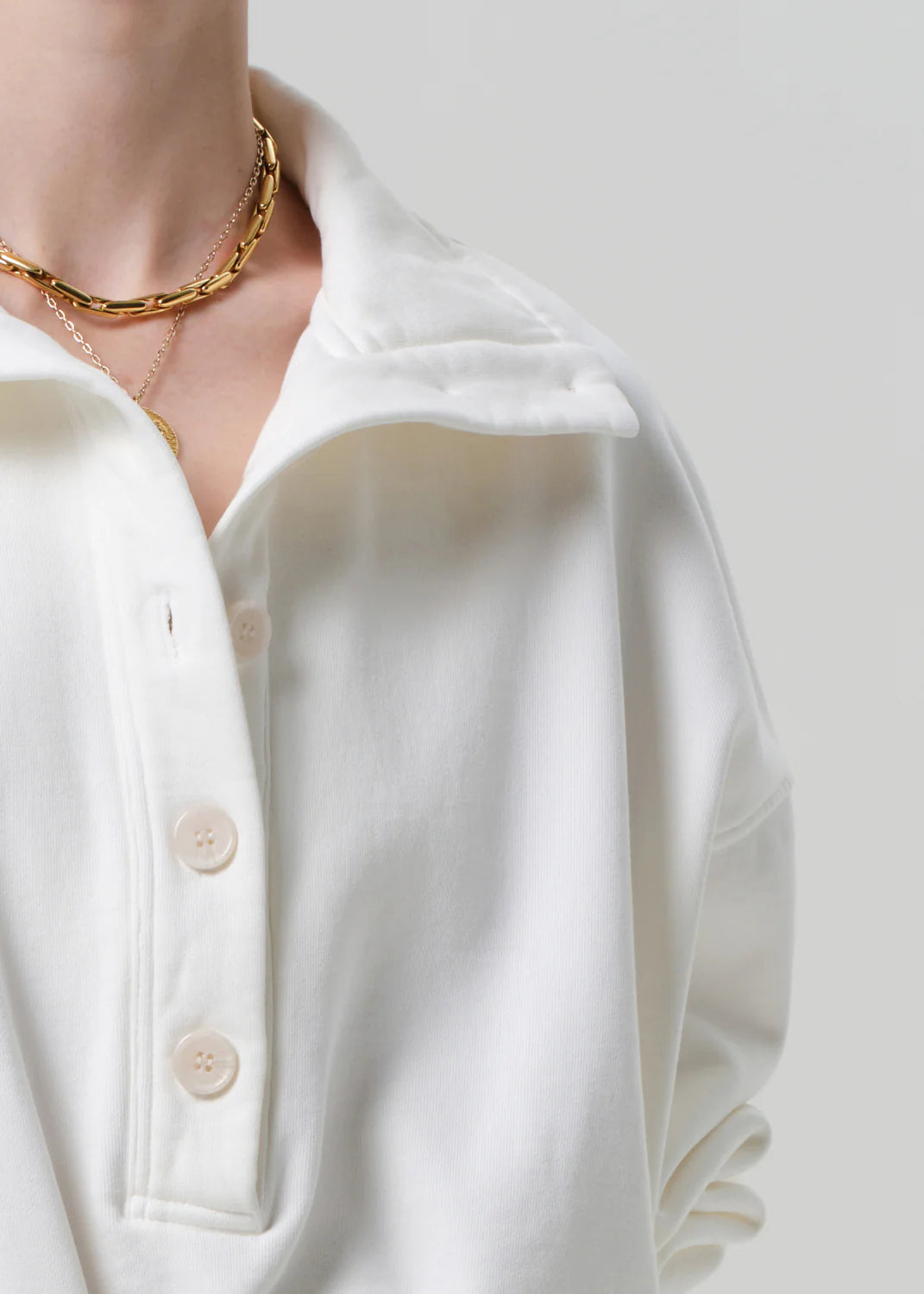 Close-up of a person wearing a white shirt with buttons and a gold necklace on a plain background