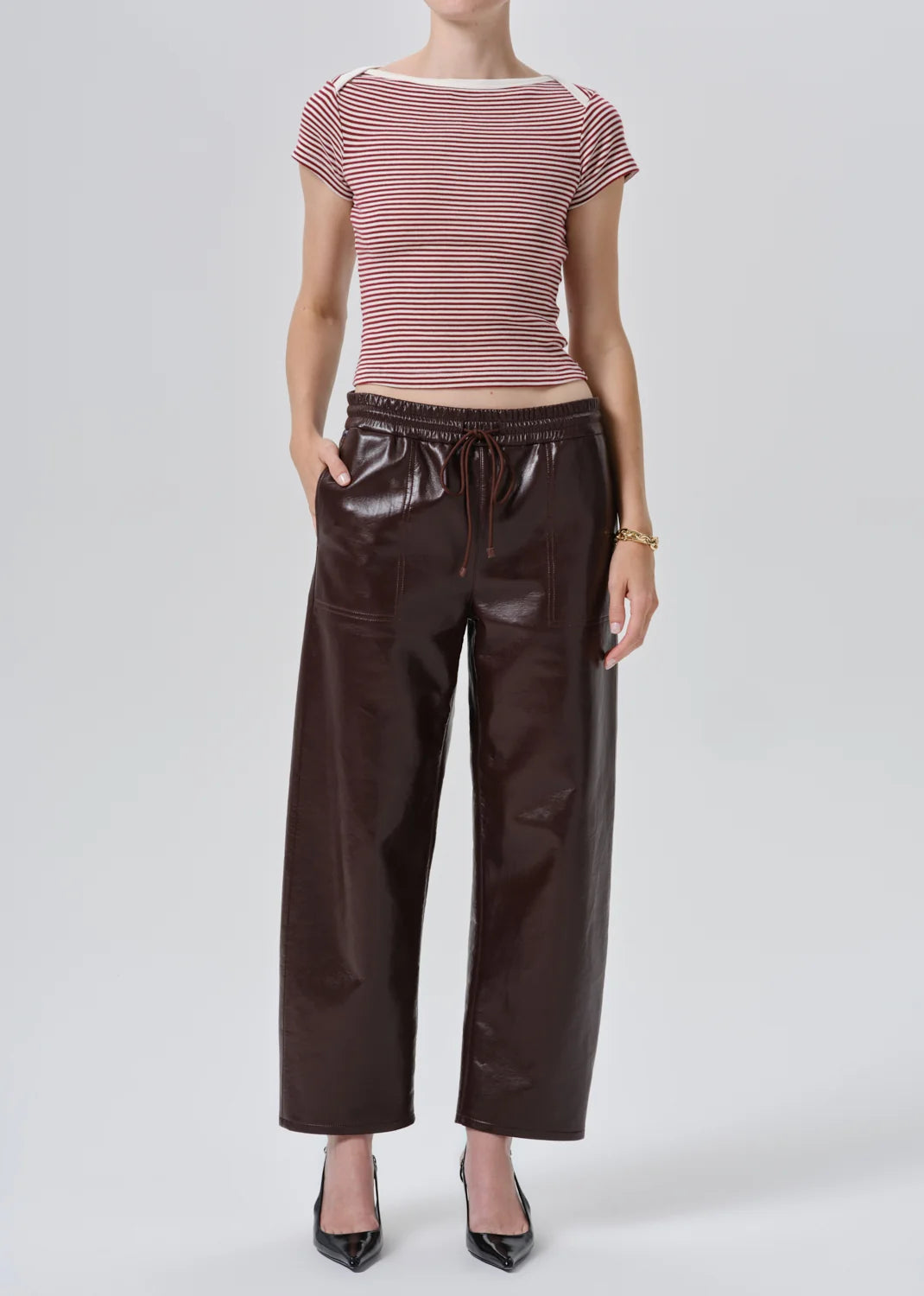 A model wearing a pink and white striped short-sleeve top with dark brown faux-leather wide-leg pants and black pointed-toe heels, photographed against a light gray studio background.