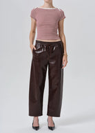 A model wearing a pink and white striped short-sleeve top with dark brown faux-leather wide-leg pants and black pointed-toe heels, photographed against a light gray studio background.