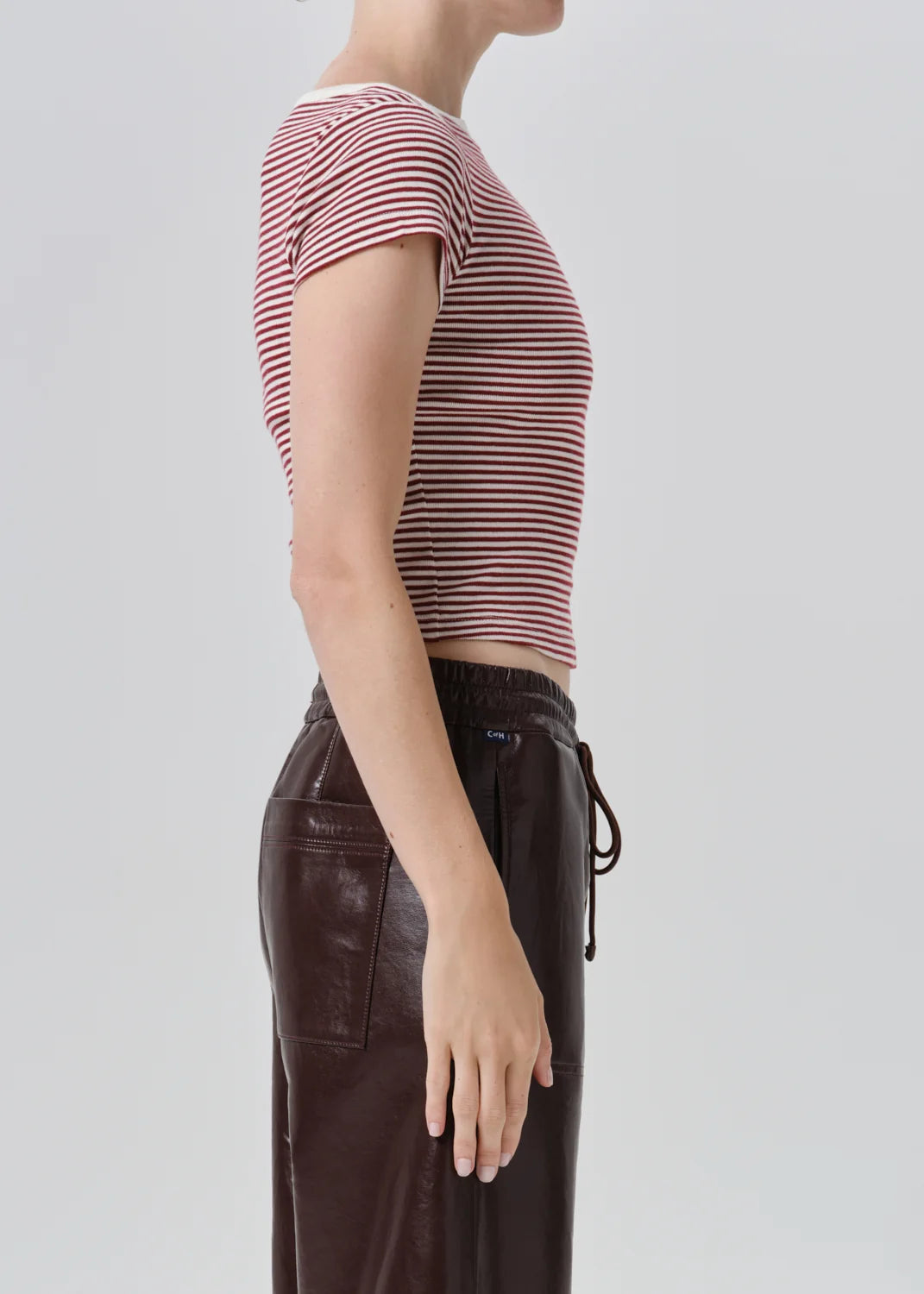 Side profile of a model wearing a short-sleeve striped crop top in red and white, paired with dark brown pants, against a light gray studio background.