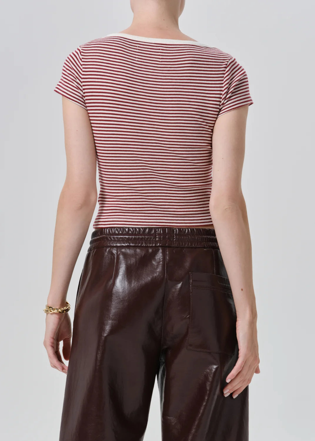 Rear view of a model wearing a short-sleeve crew-neck top with thin red and white horizontal stripes, paired with dark brown faux-leather pants, against a light gray studio background.