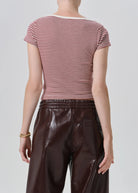 Rear view of a model wearing a short-sleeve crew-neck top with thin red and white horizontal stripes, paired with dark brown faux-leather pants, against a light gray studio background.