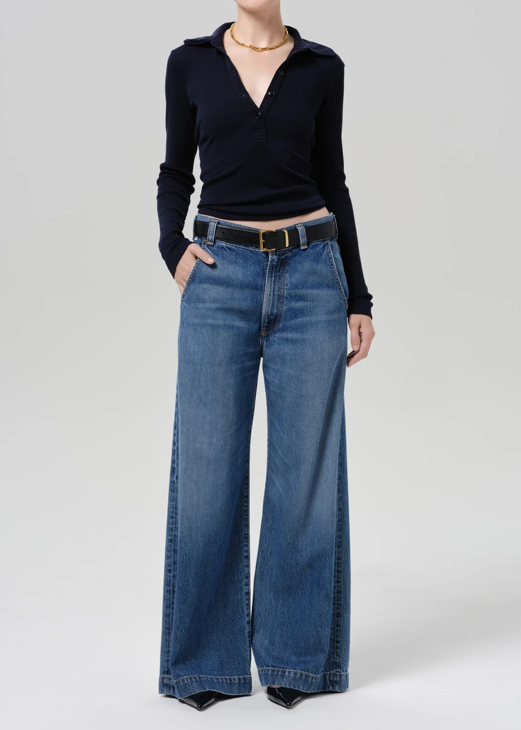A model wearing a black long-sleeve wrap top and high-waisted, wide-leg blue denim jeans with a black belt, photographed against a neutral light gray studio background.