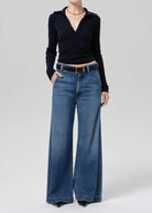 A model wearing a black long-sleeve wrap top and high-waisted, wide-leg blue denim jeans with a black belt, photographed against a neutral light gray studio background.