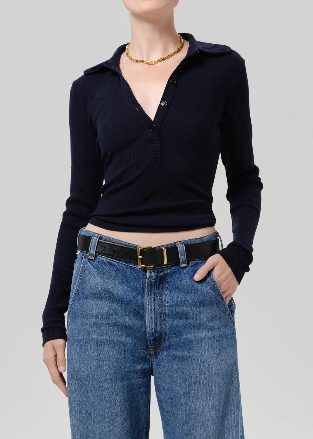 A model wearing a dark navy long-sleeve polo shirt tucked into blue jeans with a black belt, shown from the neck down against a solid gray studio background.