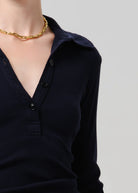 Close-up of a person wearing a dark navy ribbed long-sleeve henley top with a collar and black buttons, paired with a chunky gold chain necklace, against a light gray background.