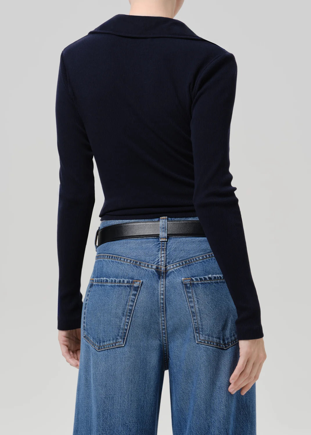 A model shown from the back wearing a dark navy long-sleeve knit top tucked into blue jeans with a black belt, against a solid light gray background.