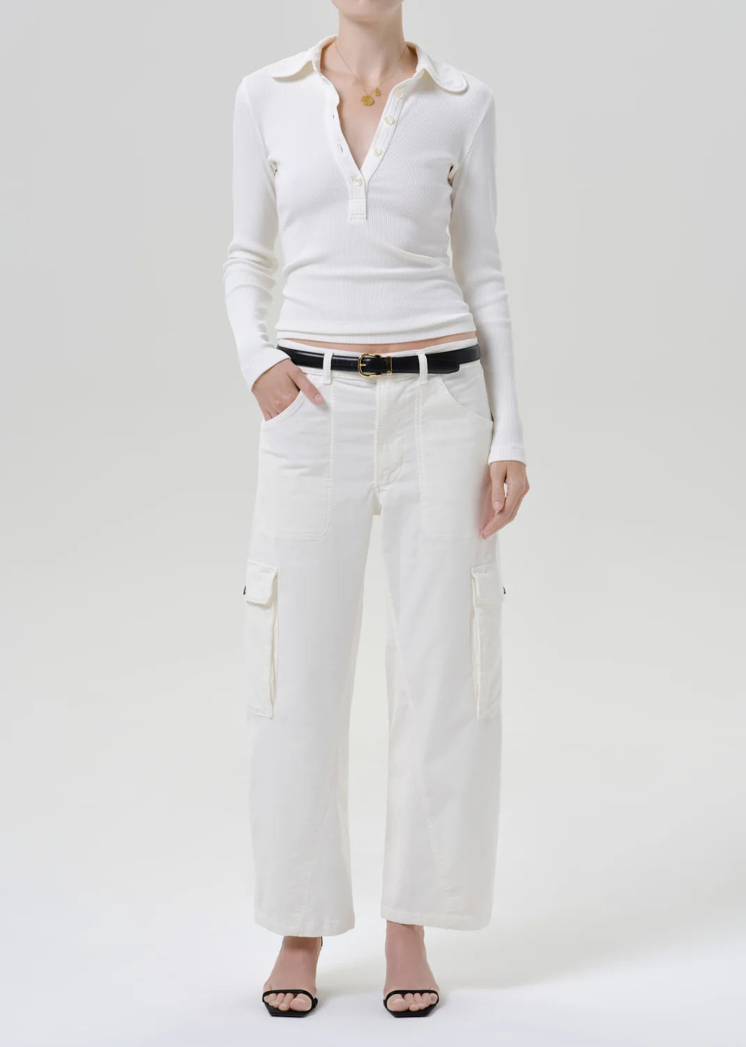 A model wearing a white long-sleeve collared knit top, white wide-leg cargo pants, a black belt, and black sandals, photographed against a seamless light gray studio background.