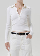 A model wearing a white ribbed long-sleeve polo shirt with a buttoned collar and chest pockets, paired with white pants and a black belt, against a light gray studio background.
